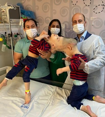 An image of Arthur and Bernardo, craniopagus twins, before their separation surgery. They are being held up by their parents and a doctor, and are on a hospital bed.