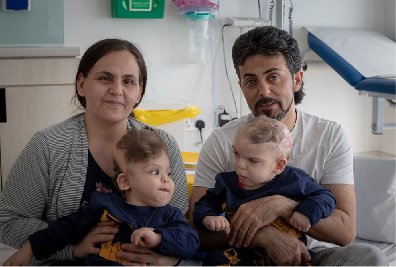The parents of Yigit and Derman hold their sons post-surgery, and look at the camera. Hospital equipment is in the background.